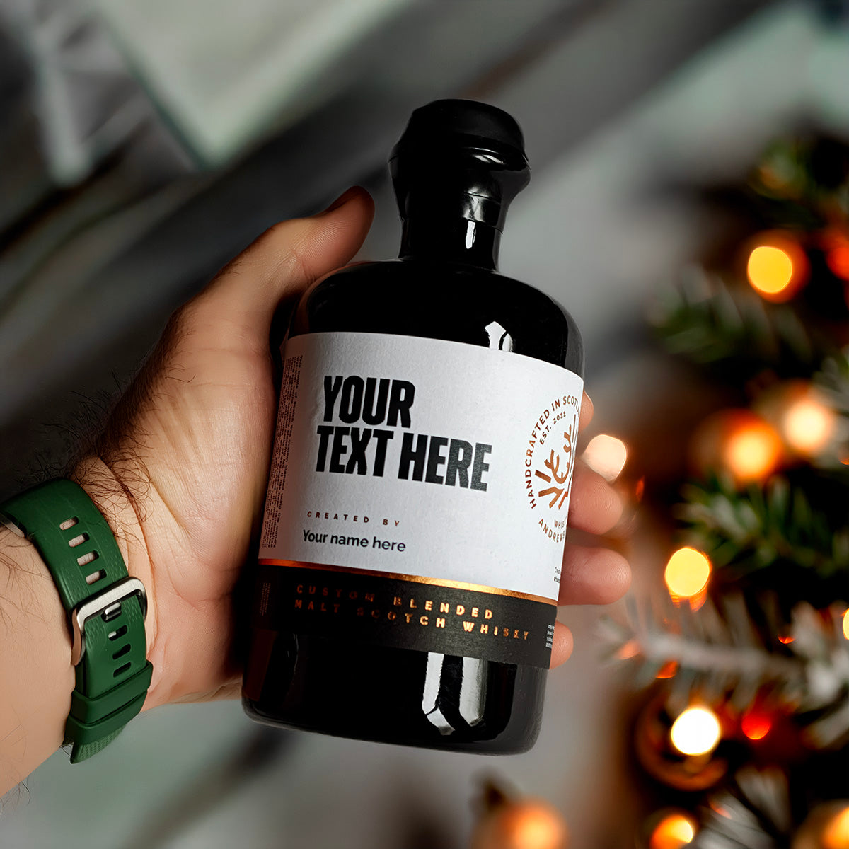 Hand holding a personalised round black whisky bottle in front of a lit Christmas tree