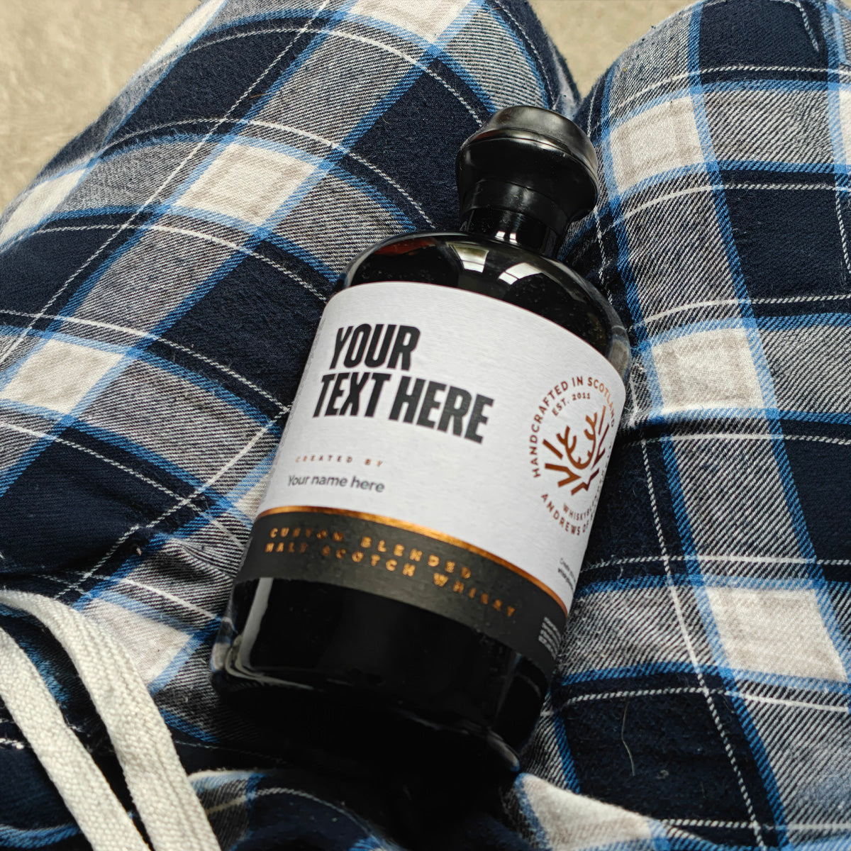 Personalised round black whisky bottle with a "Your text here" label resting on tartan pyjamas